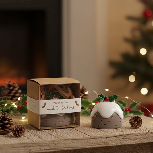 A small ceramic pudding-shaped ornament sits beside its kraft paper gift box on a wooden surface, with a blurred Christmas tree, lights, pinecones, and fireplace in the background.