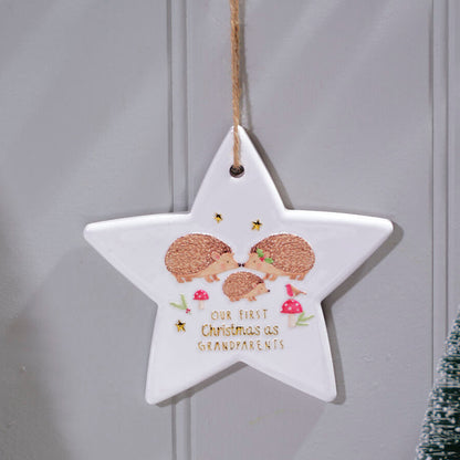 A white star-shaped Christmas hanging ornament with a natural jute hanger, featuring an illustration of three hedgehogs and the text 'Our First Christmas as Grandparents' in gold lettering. The ornament is displayed against a gray paneled wall with decorative miniature trees.