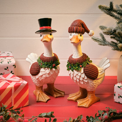Two Christmas duck figurines decorated for the holiday, standing on a red surface with wrapped gifts and evergreen branches around them. One goose wears a black top hat with a red band, and the other wears a brown knit Santa hat.