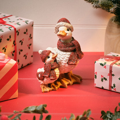 A Christmas-themed decorative figurine of a mother duck and duckling, all wearing knit red hats and scarves, posed on a red tabletop with wrapped holiday gifts in the background.