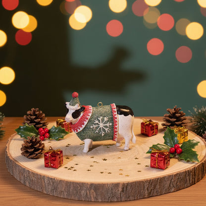 A small decorative cow figurine wearing a green Christmas sweater with white snowflakes and a red patterned collar, standing on a round wood slice coaster on a wooden table.