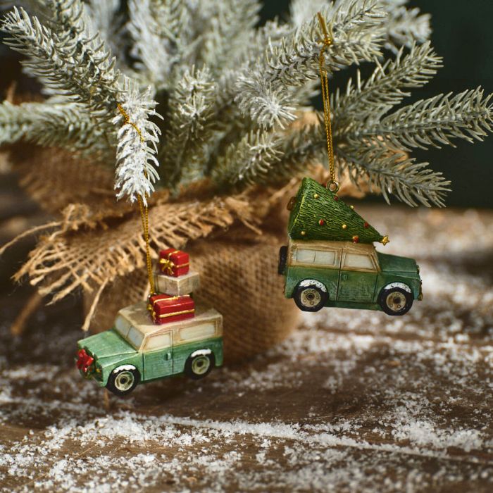 Two resin holiday ornaments featuring green vehicles, one with a red present on the roof and the other with a Christmas tree on the top, attached to a beige and brown burlap ribbon against a wooden background with snow.