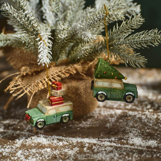 Two resin holiday ornaments featuring green vehicles, one with a red present on the roof and the other with a Christmas tree on the top, attached to a beige and brown burlap ribbon against a wooden background with snow.