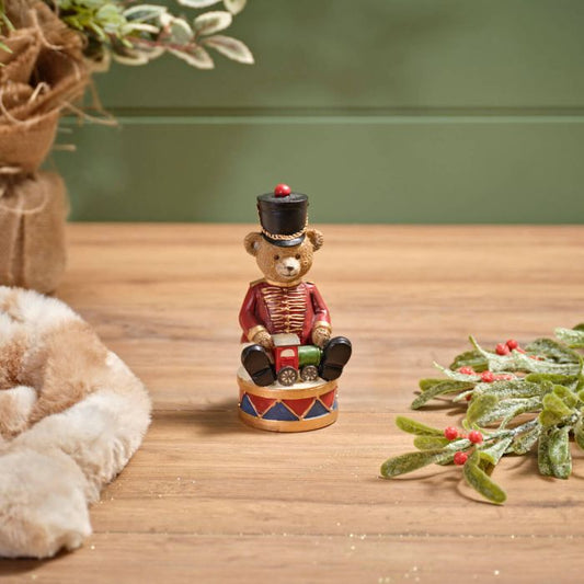 A resin ornament featuring a bear dressed as a toy soldier sitting on a drum, with a festive background.
