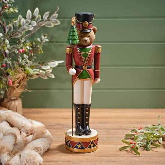 A Christmas-themed bear nutcracker standing on a drum, dressed in festive attire, with a Christmas tree in its hand, placed on a table with holiday decorations in the background.