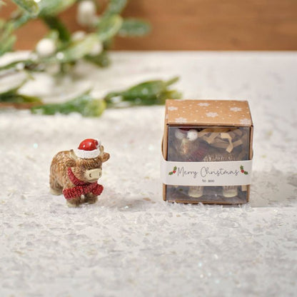 A small resin Highland cow keepsake ornament wearing a Santa hat, positioned next to its packaging which has a 'Merry Christmas' label.