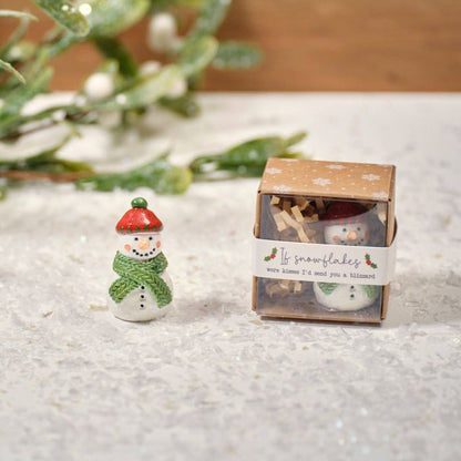 Small snowman figurine next to its packaging on a textured surface with greenery in the background