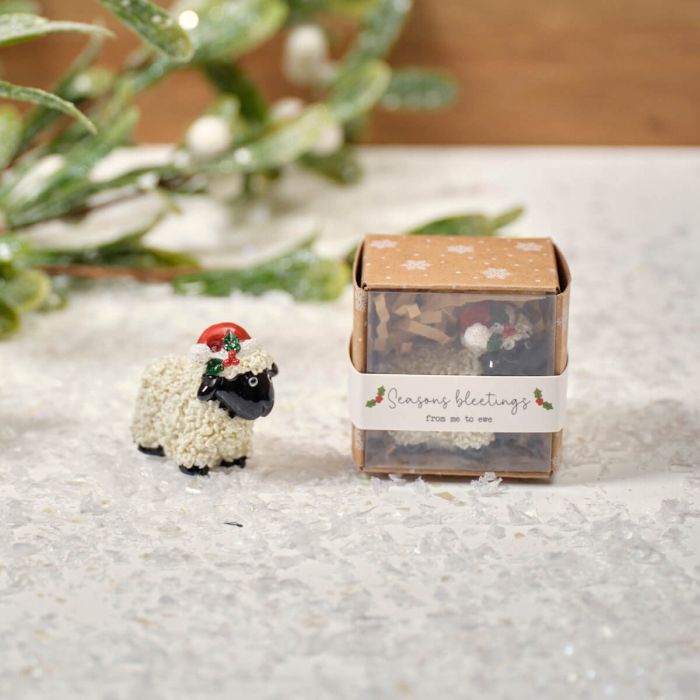 A small resin sheep keepsake ornament with a festive decoration on its head, displayed next to its packaging box with 'Season's greetings' on it, placed on a surface with artificial greenery in the background.
