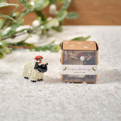 A small resin sheep keepsake ornament with a festive decoration on its head, displayed next to its packaging box with 'Season's greetings' on it, placed on a surface with artificial greenery in the background.