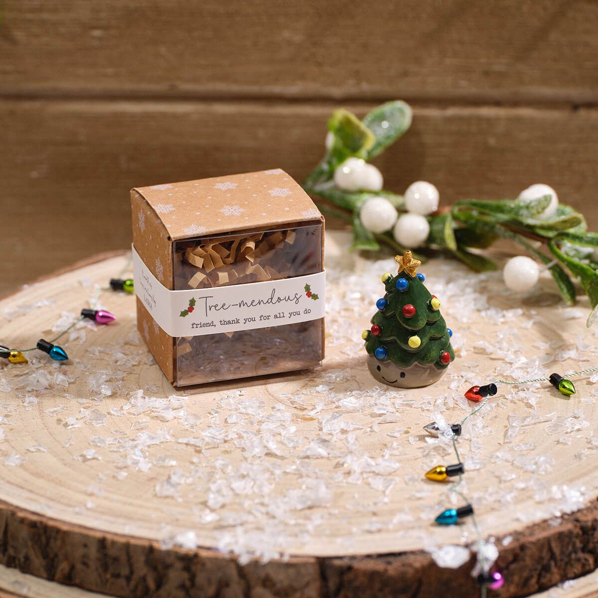 A small kraft paper gift box with a white label reading 'tree-mendous' sits on a rustic wood slice, styled with a miniature resin Christmas tree, white berries, string lights, and white flakes against a wooden background.
