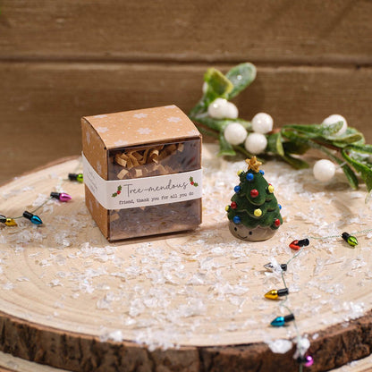 A small kraft paper gift box with a white label reading 'tree-mendous' sits on a rustic wood slice, styled with a miniature resin Christmas tree, white berries, string lights, and white flakes against a wooden background.