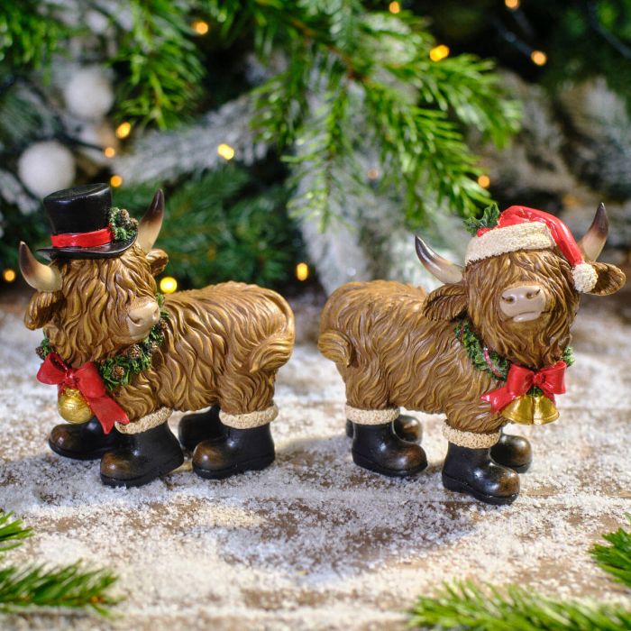 Two decorative Highland cow figurines with festive attire on a snowy surface with Christmas tree in the background.