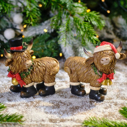 Two decorative Highland cow figurines with festive attire on a snowy surface with Christmas tree in the background.