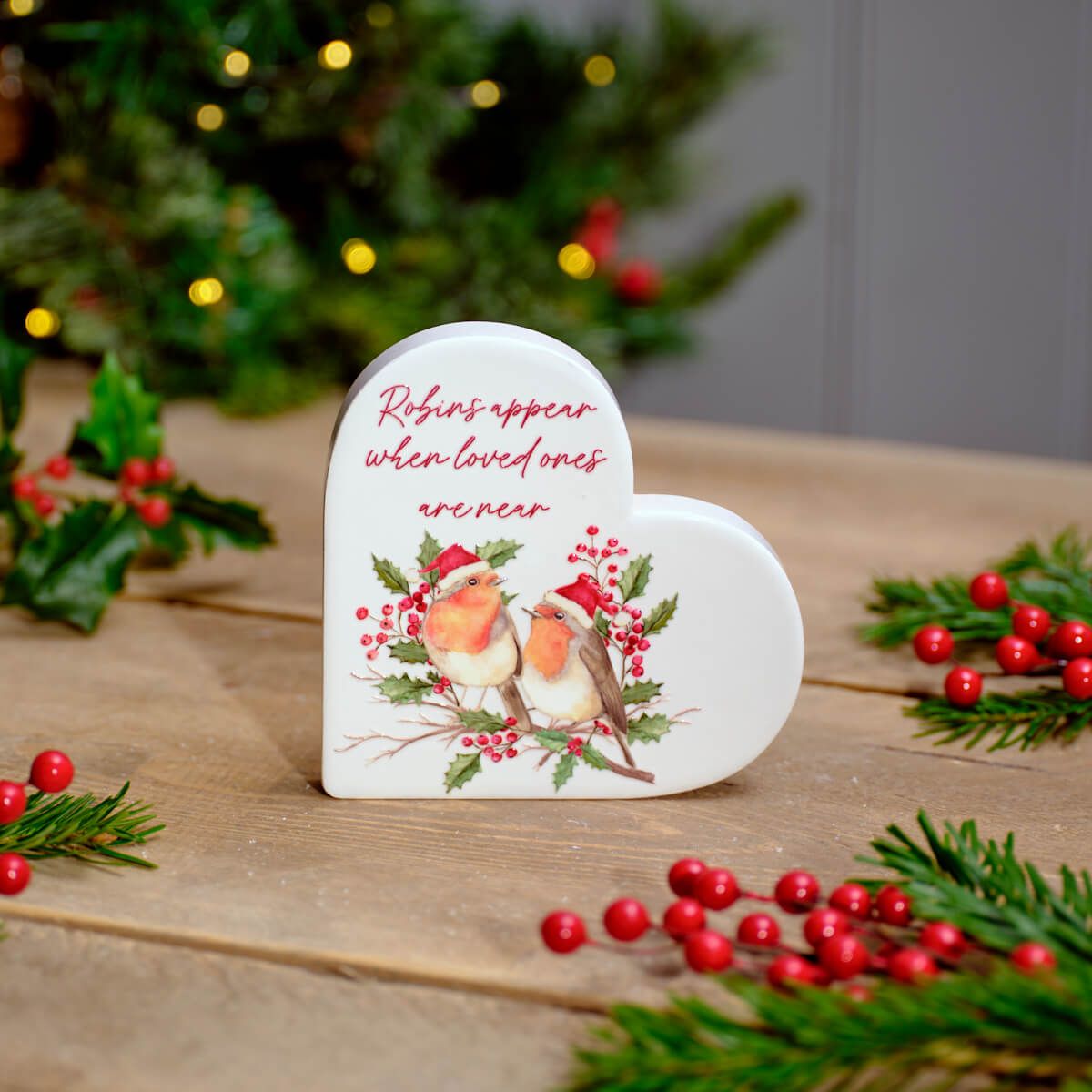 A ceramic heart-shaped block with a Christmas-themed design, featuring two robins and festive berries, placed on a wooden surface with holiday decorations in the background.