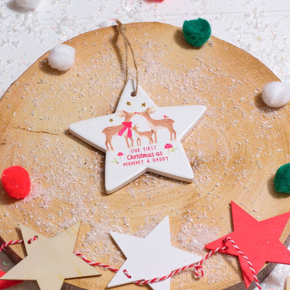 A white ceramic Christmas ornament shaped like a star hangs from a string on a round wooden slice. The ornament is printed with a reindeer illustration and the text 'Our First Christmas is Mummy & Daddy'.