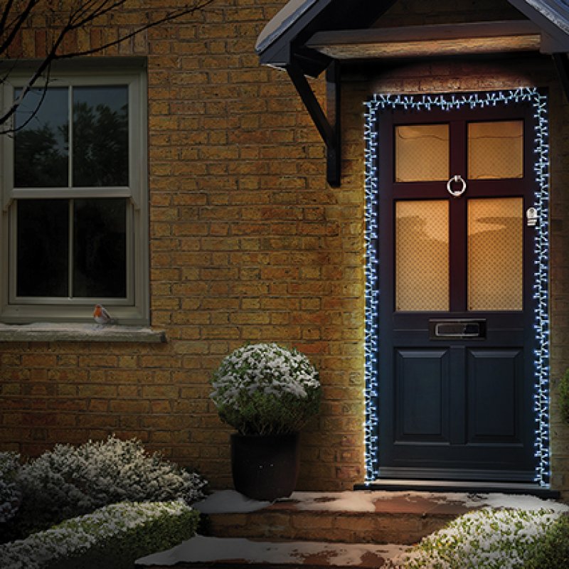 A front door of a brick house decorated with white string lights around the door frame and door panels, with a small snowball in a planter on the steps.