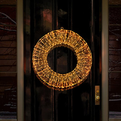 A circular holiday wreath decorated with warm white string lights, hanging on a dark front door at night.