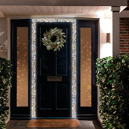 Outdoor Christmas string lights draped around a front door, with a green wreath hanging on the door, set in a real residential entryway at night.