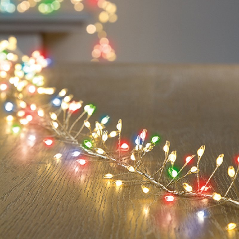 Multicolor Christmas string lights draped across a wooden tabletop, with warm white bulbs and scattered red, green, and blue lights in the foreground and soft bokeh lights in the background.
