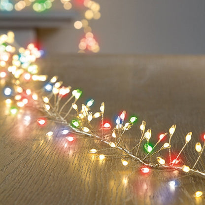 Multicolor Christmas string lights draped across a wooden tabletop, with warm white bulbs and scattered red, green, and blue lights in the foreground and soft bokeh lights in the background.