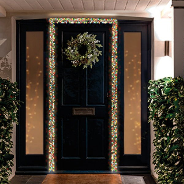 A front door at night decorated with a green wreath and illuminated holiday garlands framing the door, shown on a house porch with brick walls and shrubs.