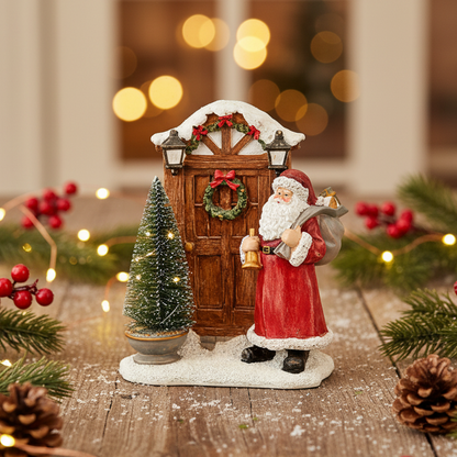 A festive Christmas figurine depicting Santa Claus in a red suit with white trim, standing next to a snow-covered wooden door with a wreath, bell, and lantern, with a small decorated tree on a snowy base.