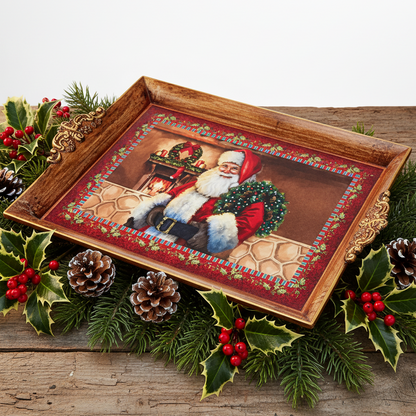 A rectangular wooden serving tray with ornate gold-toned handles and a festive Christmas print of Santa Claus, styled on a rustic wooden surface with pine branches, holly, pinecones, and red berries.
