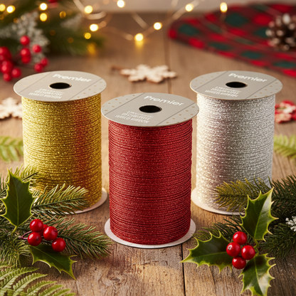 Three spools of Premier Christmas ribbon in gold, red, and silver, styled with holly leaves and red berries on a rustic wooden table with festive string lights in the background.