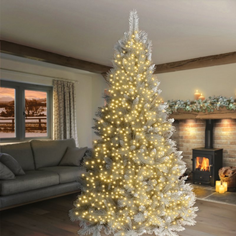 A tall artificial Christmas tree with white faux snow and warm white string lights, standing in a cosy living room with a fireplace and sofa in the background.