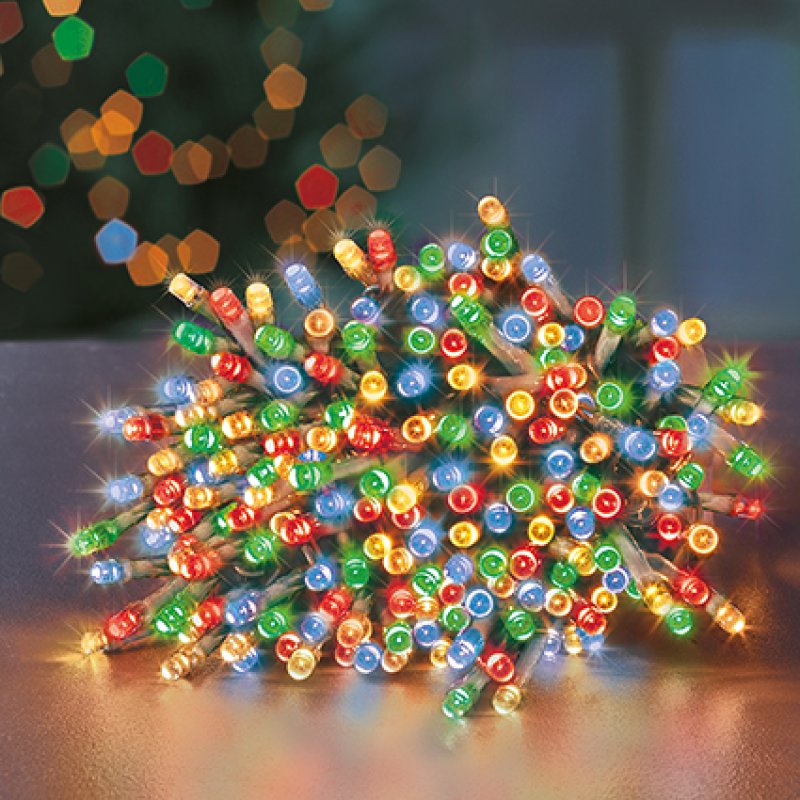 Multicolor Christmas string lights with many small round bulbs in red, green, blue, yellow, and white, clustered on a tabletop with a blurred holiday background.