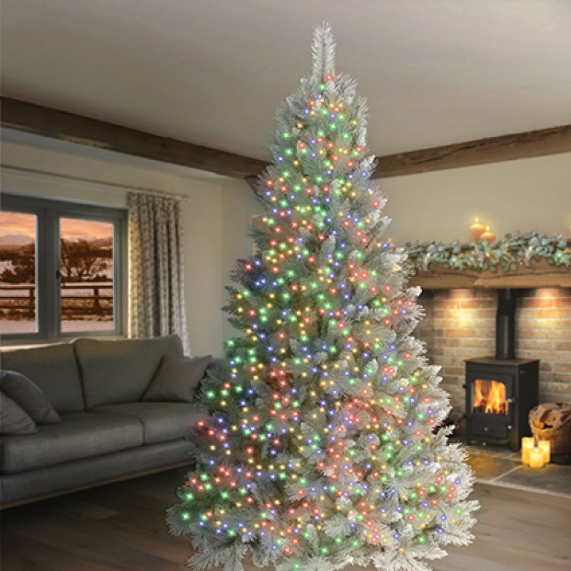 A tall, flocked artificial Christmas tree decorated with multicolored string lights, set in a cosy living room with a fireplace and sofa in the background.