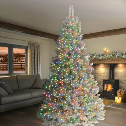 A tall, flocked artificial Christmas tree decorated with multicolored string lights, set in a cosy living room with a fireplace and sofa in the background.