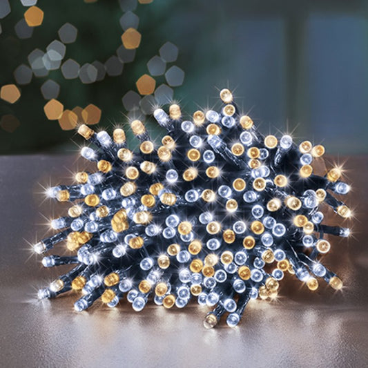 A cluster of decorative string lights with small round bulbs, glowing in white and warm white tones, arranged on a reflective surface with a blurred festive background.