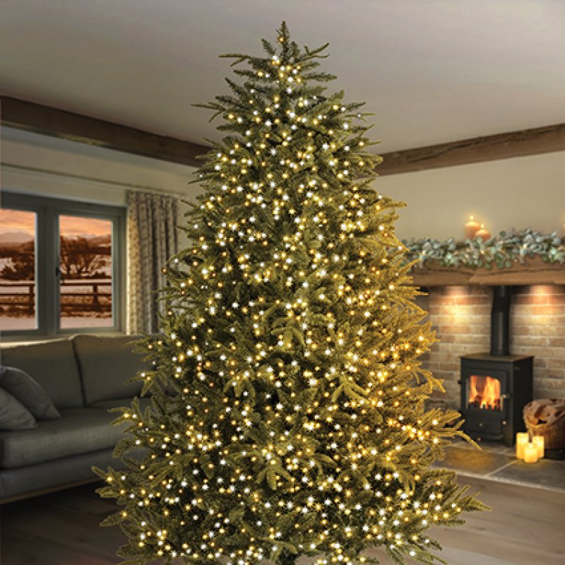 A tall artificial Christmas tree with warm white string lights, set in a cozy living room with a fireplace and sofa in the background.