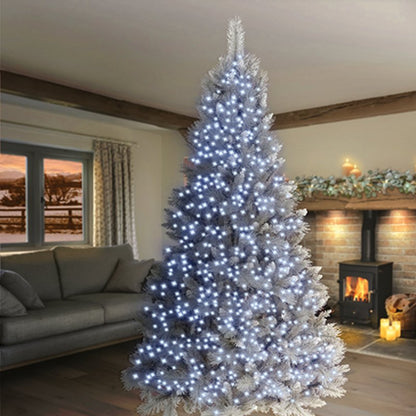 A tall artificial Christmas tree with white faux snow and glowing white lights, placed in a cozy living room with a fireplace and sofa in the background.