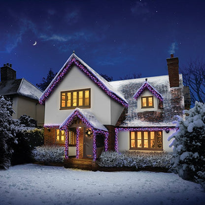 A snow-covered house at night decorated with rainbow string lights along the roofline, porch, and windows, with warm yellow lights glowing from the lit windows under a dark blue starry sky.