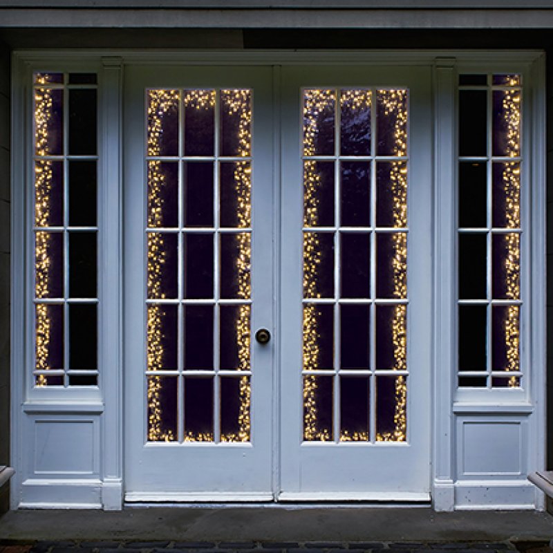 White French door double doors with tall grid-style glass panes, decorated with warm white string lights framing the glass panels, shown against a dark evening background.
