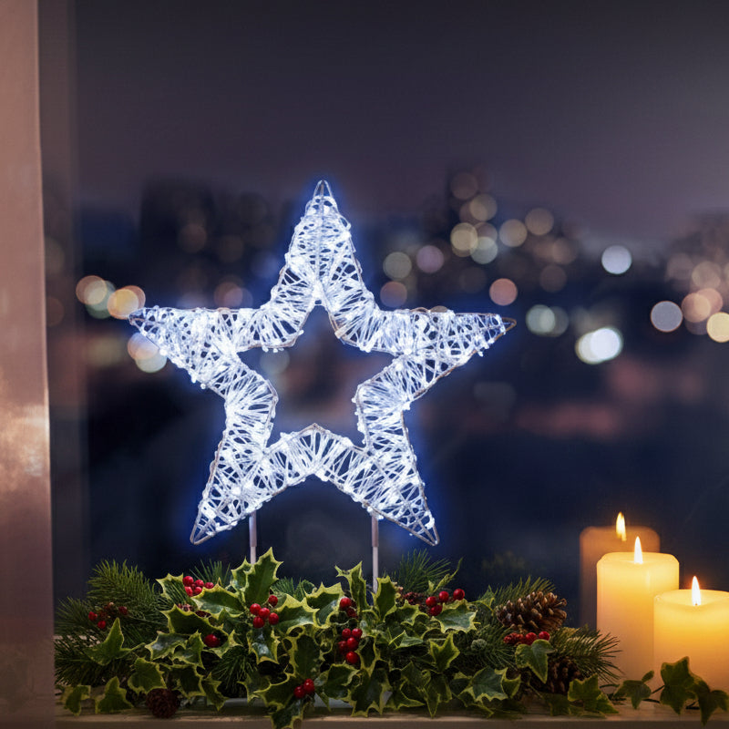 A glowing star-shaped decorative light on a small stand, placed on a windowsill at night with a blurred city view outside and warm window light to the left.