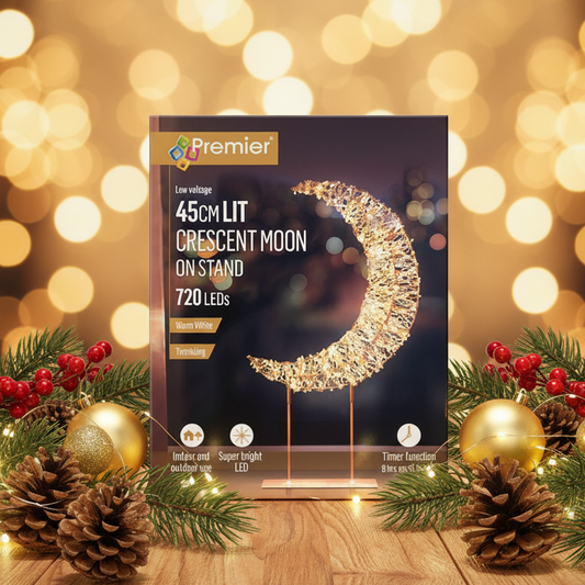 A boxed LED crescent moon lamp stand by Premier, shown upright on a wooden surface with festive pine branches, pinecones, red berries, and gold ornaments against a warm bokeh holiday background. The package text reads 45cm, 45cm lit, crescent moon on stand, 720 LEDs, warm white.