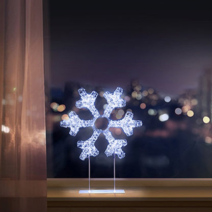 A glowing snowflake LED light on a clear acrylic stand, displayed on a windowsill with a blurred city night view outside.