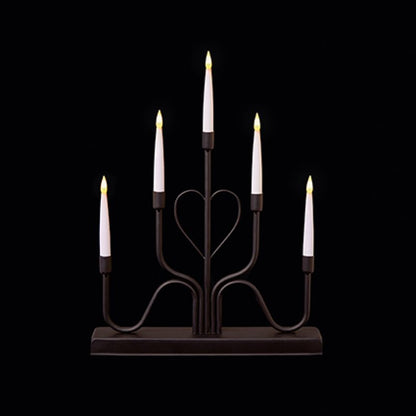 A six-light candle-style candelabra with a dark metal frame and five white faux candles, each lit with a warm yellow flame, set against a solid black background.