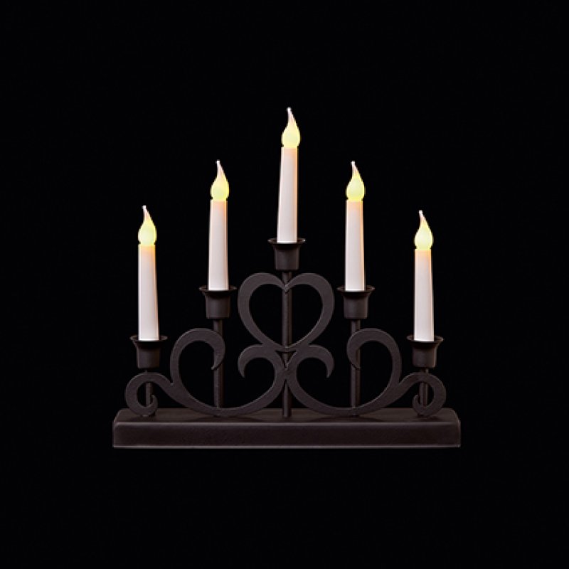A five-light candle-style table lamp with a dark metal scrollwork base and five white faux candles, each lit with a warm yellow flame effect, shown against a solid black background.