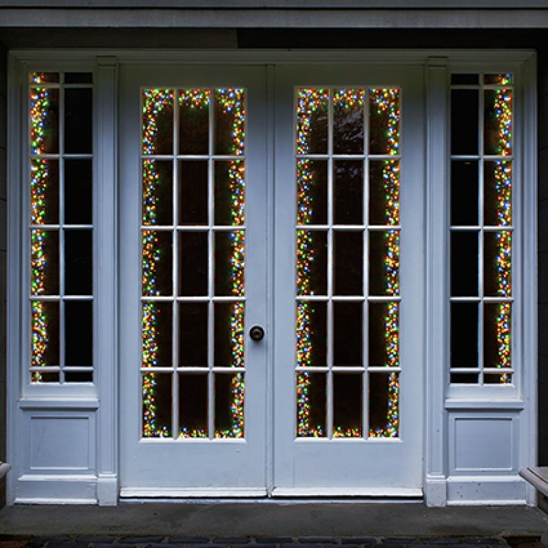 White French door windows decorated with vertical strands of multicolor Christmas string lights, set against a dark background.