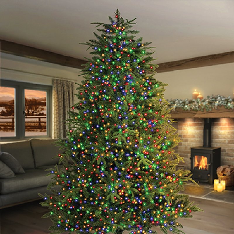 A tall artificial Christmas tree decorated with multicolored string lights, standing in a cosy living room with a fireplace, sofa, and window visible in the background.