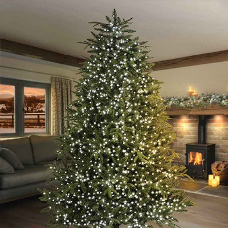 A tall artificial Christmas tree decorated with glowing white string lights, set in a cosy living room with a fireplace, sofa, and warm ambient lighting.