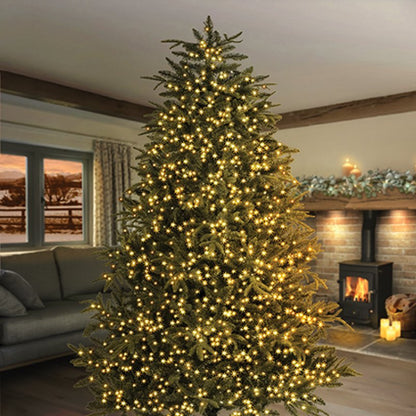 A tall artificial Christmas tree covered in warm white string lights, standing in a cozy living room with a fireplace, sofa, and window in the background.