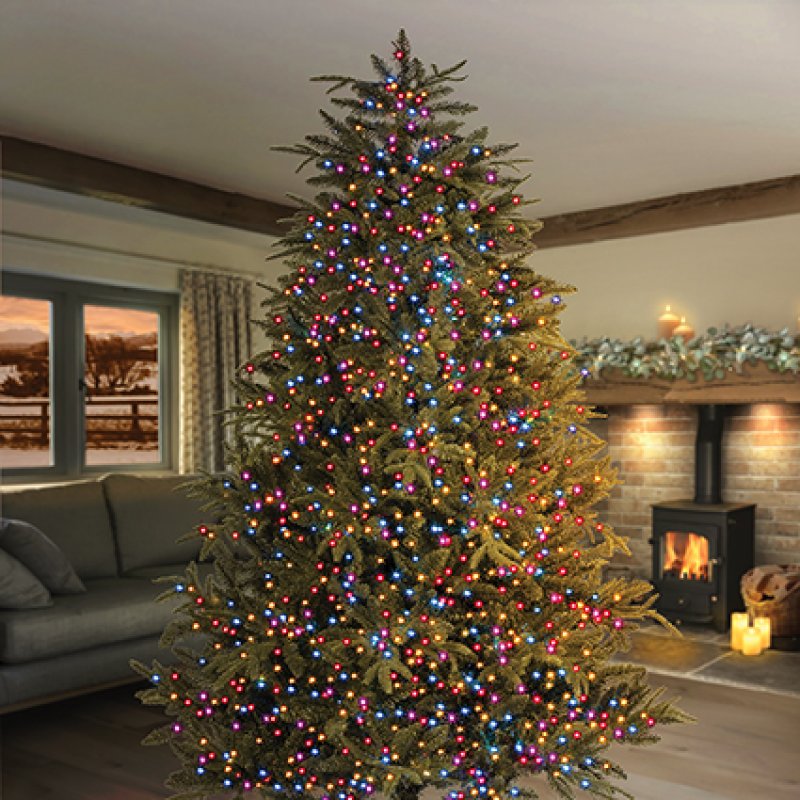 A tall artificial Christmas tree in a living room, decorated with rainbow colour led string lights, with a fireplace and sofa in the background.