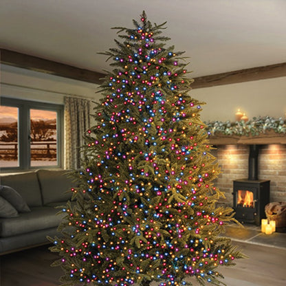 A tall artificial Christmas tree in a living room, decorated with rainbow colour led string lights, with a fireplace and sofa in the background.