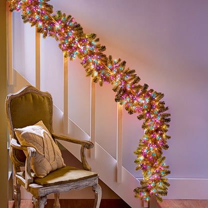 A festive evergreen garland with rainbow Christmas lights draped over a staircase railing next to an antique-style upholstered armchair.