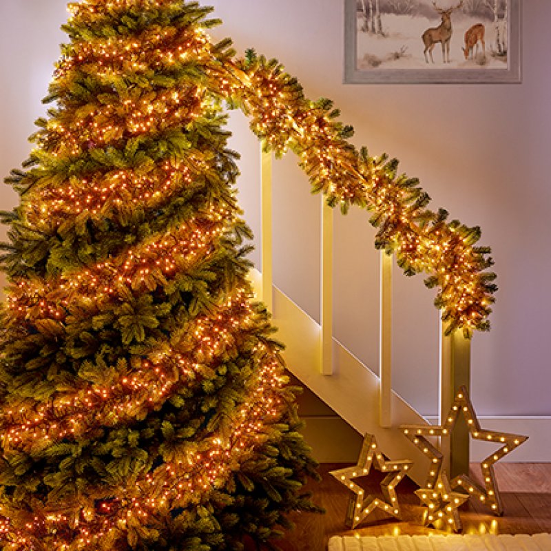 A decorated Christmas tree and staircase railing with vintage gold string lights in a home interior, with illuminated star ornaments on the floor and a framed deer picture on the wall.